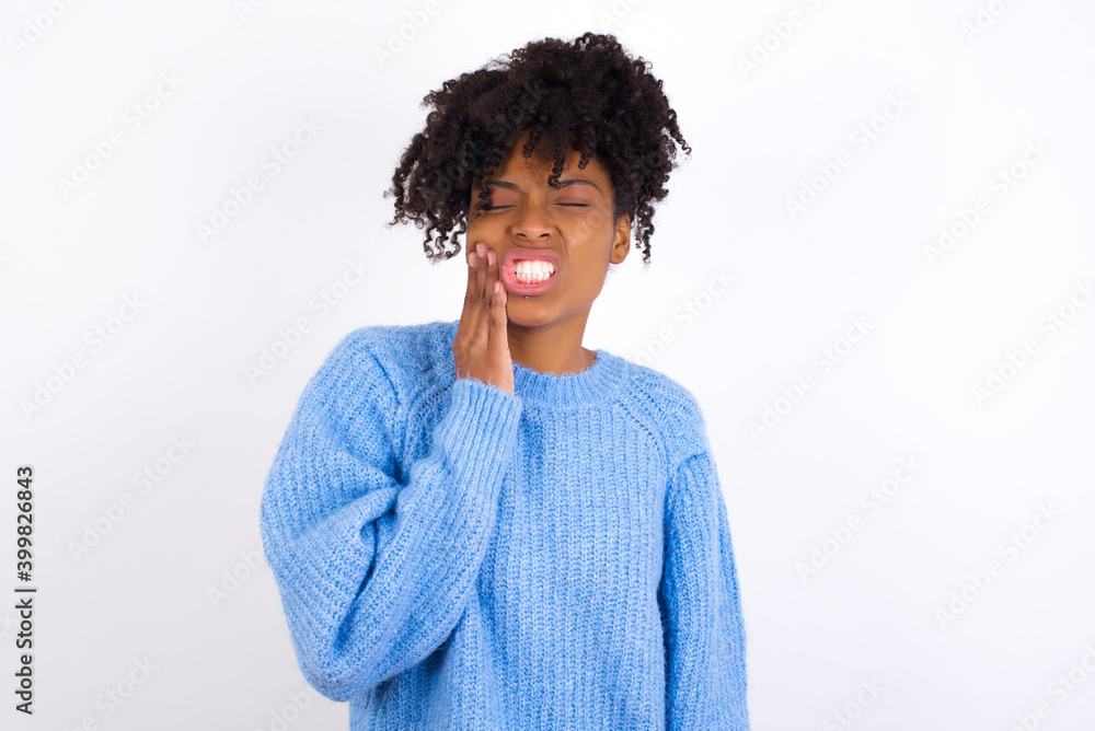 Young beautiful African American woman wearing blue knitted sweater against white wall touching mouth with hand with painful expression because of toothache or dental illness on teeth. Young beautiful African American woman wearing blue knitted sweater against white wall touching mouth with hand with painful expression because of toothache or dental illness on teeth.