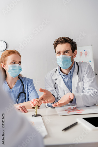 Doctor in medical mask talking to colleagues while sitting at workplace with papers and brain anatomical model on blurred foreground