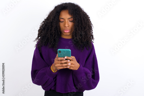 Excited Young beautiful African American woman wearing knitted sweater against white wall winking and eye hold smart phone use read social network news