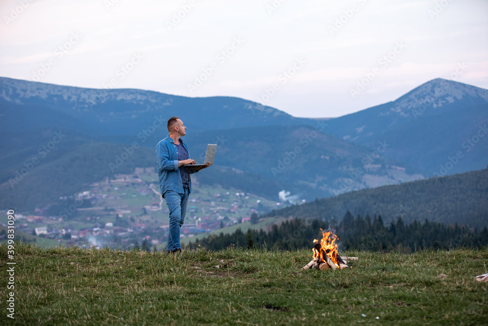 Man working outdoors with laptop standing in mountains. Concept of remote work or freelancer lifestyle. Cellular network broadband coverage. internet 5G. Hiker tourist enjoying valley view