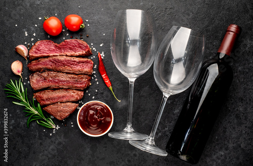 Different degrees of roasting beef steak in the shape of a heart with spices on a meat knife and bottles of red wine with glasses on a stone background. valentines day celebration concept