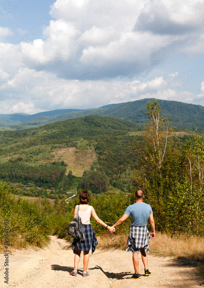 Back view of two young people holding hands standing together on mountain road. Sunny and bright day. Hiking concept. Vacation in the mountains.