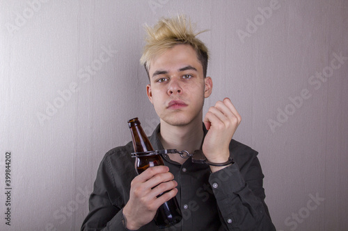 A teenager in handcuffs and with a bottle in his hand on a gray background. Concept: adolescent alcoholism, early attachment to drinking, poor heredity.