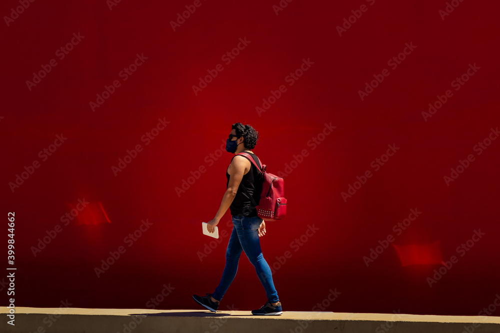 Jovem homem, caminhando na rua, usando máscara de proteção, com mochila vermelha nas costas e muro pintado de vermelho atrás.