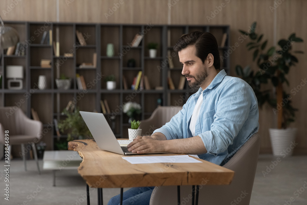 Millennial Caucasian businessman sit at desk at home office look at laptop screen working with paperwork documents. Serious young man wok on computer online at workplace, consult client or customer.