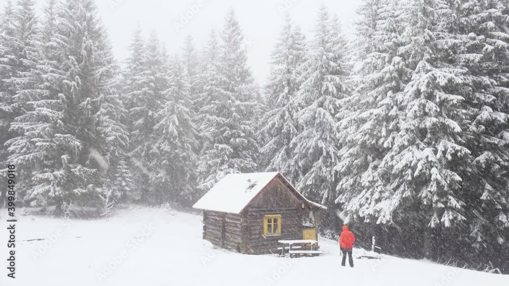 Man hiker in red jacket walking into a log wooden cabin. Snow falling on dark mountain pine forest . Christmas holidays and winter vacations concept Man hiker in red jacket walking into a log wooden cabin. Snow falling on dark mountain pine forest . Christmas holidays and winter vacations concept