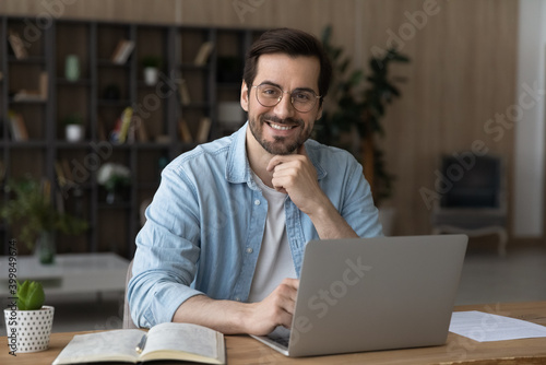 Portrait of smiling millennial Caucasian male employee in glasses sit at desk at home office working distant on laptop. Happy young 20s man in spectacles use computer, consult client customer online.