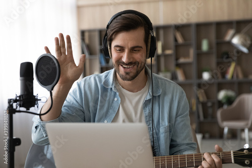 Fototapet Happy millennial male artist in headphones and guitar have online video music lesson on computer