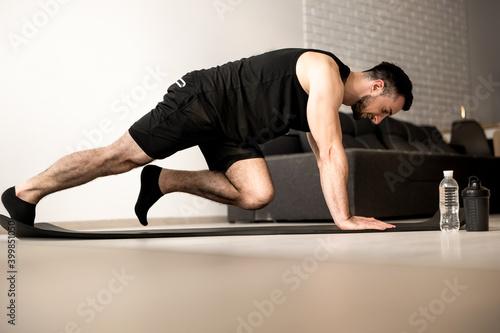 Man doing mountain climber exercises on black yoga mat. Morning workout. White modern living room on background. Plastic bottle of water. Hard workout. Training at home.
