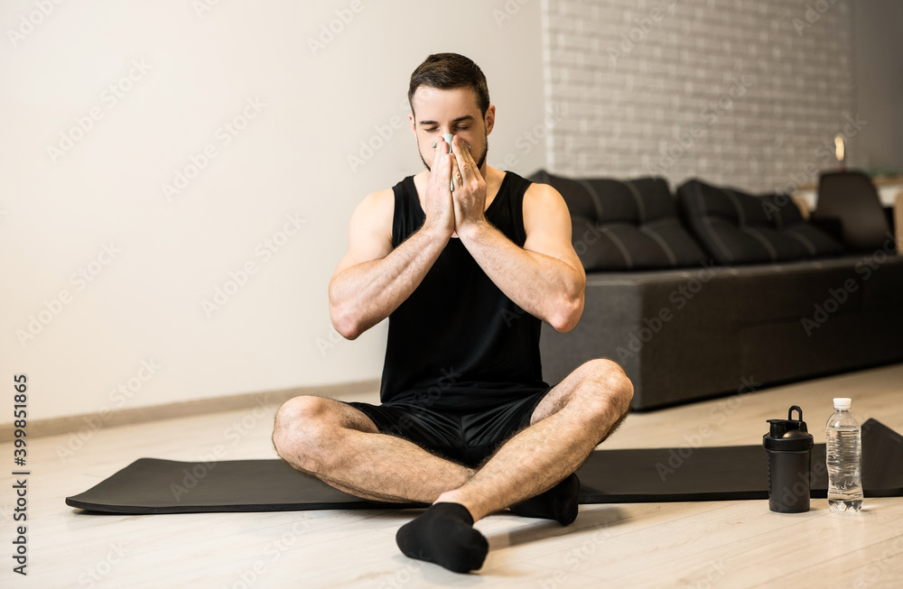 Sick man with runny nose sitting on black yoga mat. Stay home concept. Brunette young man blows his nose after workout. Practicing sports at home if you feel sick.