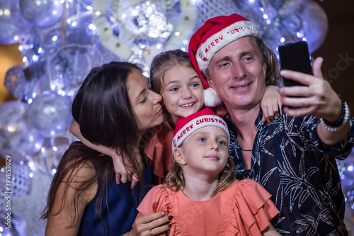 Smiling mother, father and two little daughters are making selfie with smartphone over christmas tree lights background. Family, holidays, technology and people concept