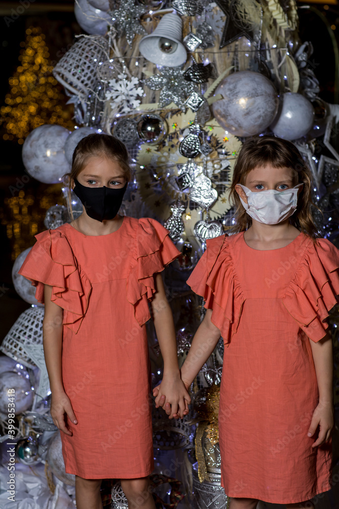 Two cute sisters standing near Christmas tree, holding hands and wearing protective face masks. Quarantine, isolation, social distance. Merry Christmas and happy holidays during pandemic
