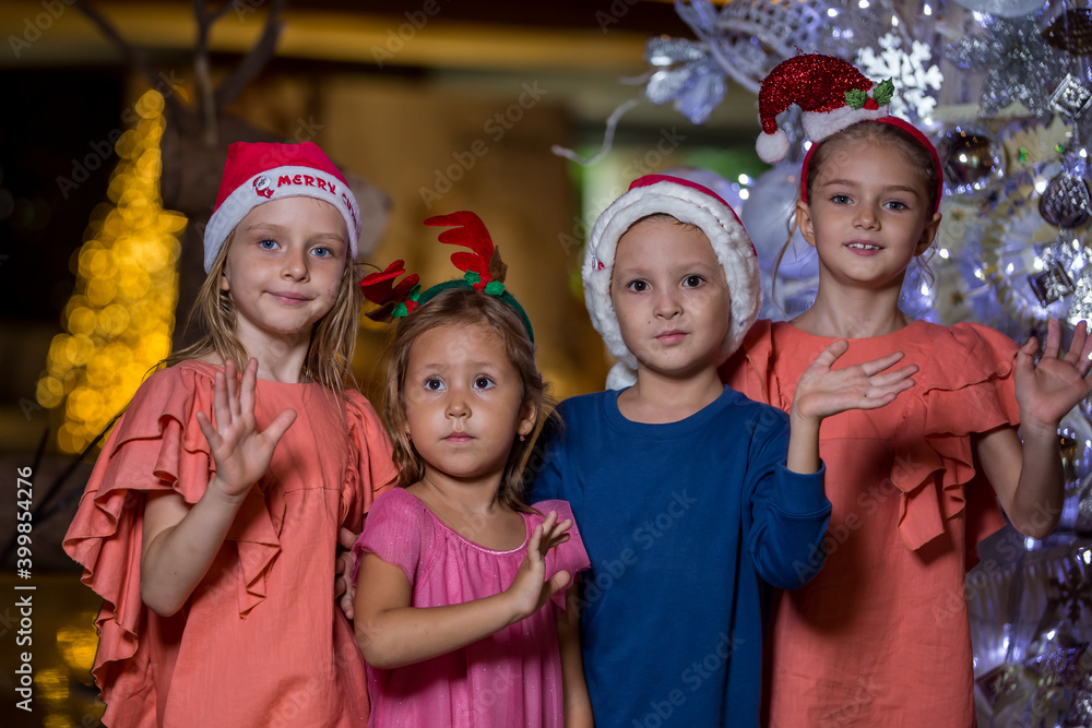 Happy cute little children wearing Santa hats standing near Christmas tree, waving and greeting. Merry Christmas and happy holidays Happy cute little children wearing Santa hats standing near Christmas tree, waving and greeting. Merry Christmas and happy holidays