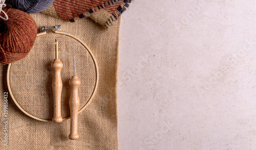 Composition with copy space of punch needle tools on a concrete surface. A jute fabric in an embroidery hoop, two different size of needles and wool balls.