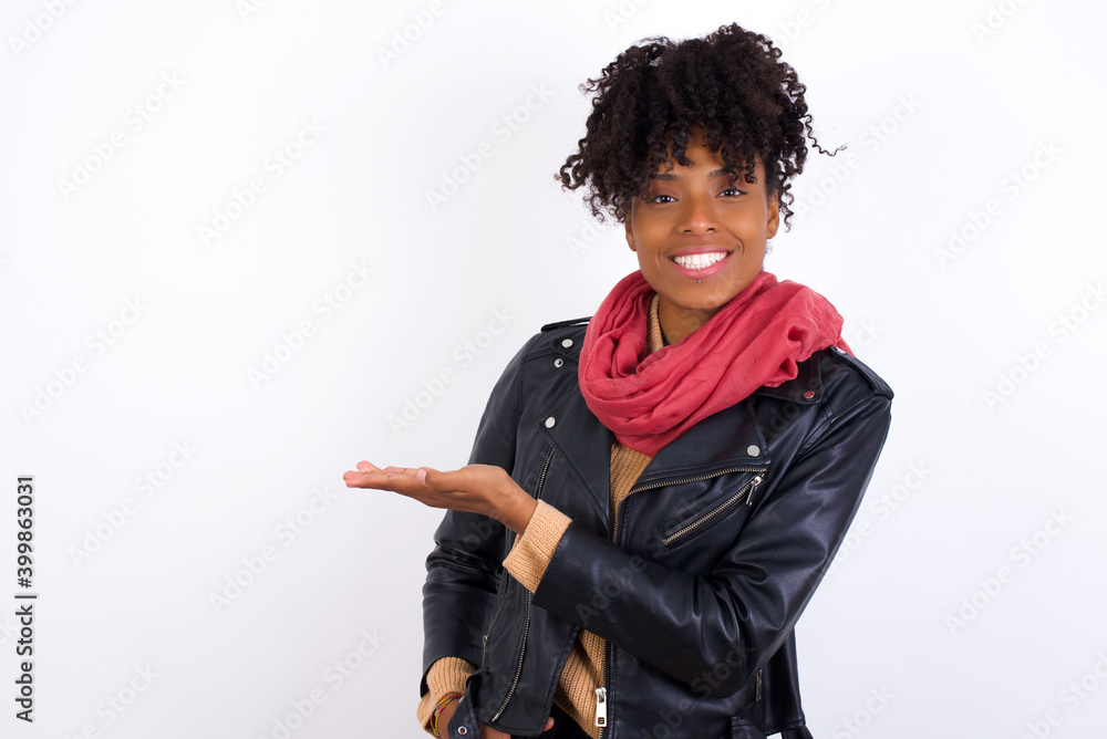 Young beautiful African American woman wearing biker jacket against white wall says: wow how exciting it is, has amazed expression, shows something on blank space with palm. Advertisement concept. Young beautiful African American woman wearing biker jacket against white wall says: wow how exciting it is, has amazed expression, shows something on blank space with palm. Advertisement concept.