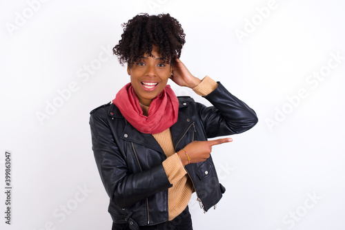 Young beautiful African American woman wearing biker jacket against white wall feeling positive has amazed expression, indicates something. One hand on head and pointing with other hand.
