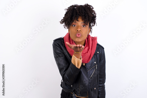 Young beautiful African American woman wearing biker jacket against white wall looking at the camera blowing a kiss with hand on air being lovely and sexy. Love expression.