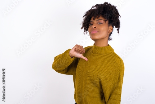 Closeup of cheerful Young beautiful African American woman wearing knitted sweater against white wall looks joyful, satisfied and confident, points at himself with thumb.
