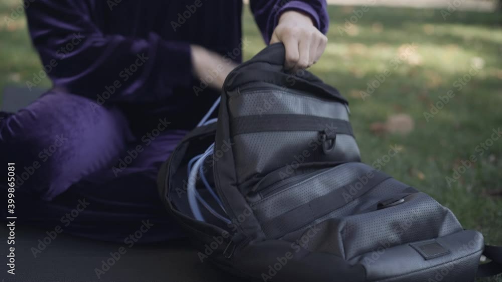 Unrecognizable sportswoman sitting on exercise mat outdoors packing and zipping backpack. Young slim Caucasian woman after morning training on green summer meadow. Unrecognizable sportswoman sitting on exercise mat outdoors packing and zipping backpack. Young slim Caucasian woman after morning training on green summer meadow.