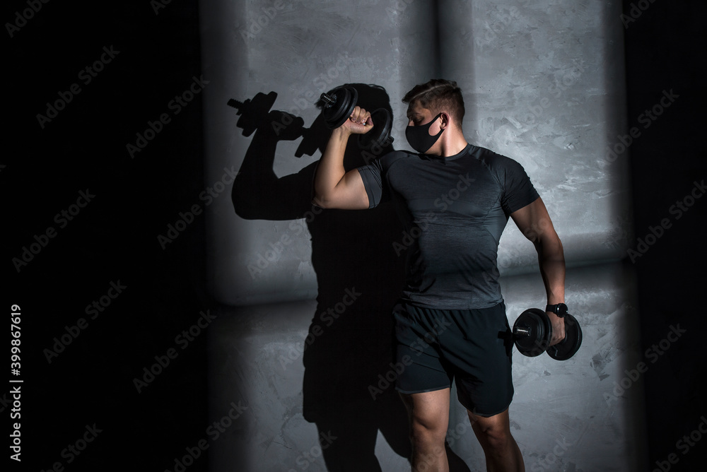 Male athlete wearing protective face mask and training with dumbbell in gym. Coronavirus world pandemic and sport theme.