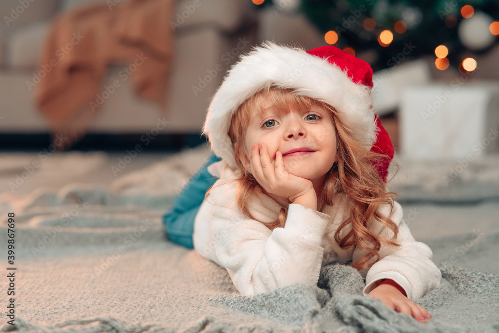 Beautiful little girl with white curly hair and Christmas hat lies in the room on the floor against the background of decorated Christmas tree and lights Beautiful little girl with white curly hair and Christmas hat lies in the room on the floor against the background of decorated Christmas tree and lights