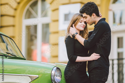 Young multiracial couple, male and female lovers heterosexual people students. Beautiful models posing standing near a retro car in the city. Dressed in black clothes