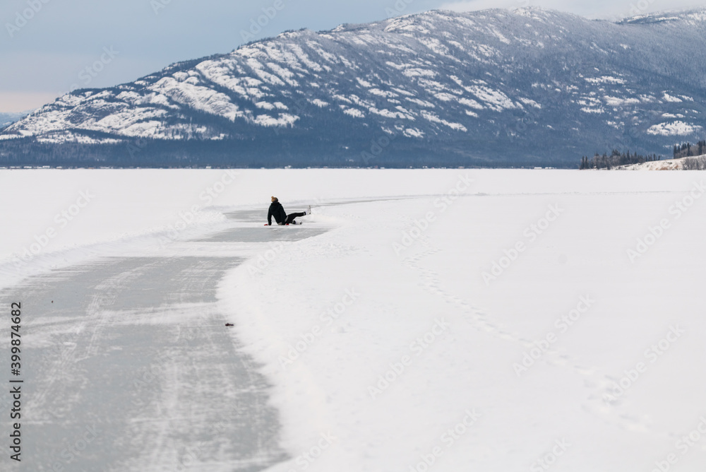 Woman fallen while skating on frozen lake in northern Canada with huge mountains in the background surrounded by snow, snow capped and glassy ice on lakes surface. 