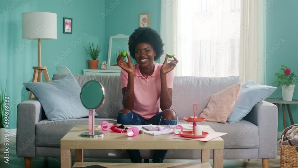 Portrait of young stylish Afro-American woman in pink T-shirt, sitting on sofa, smiling at camera, playing with lime halves, Concept of beauty and fashion, Zoom out, Slow motion. Portrait of young stylish Afro-American woman in pink T-shirt, sitting on sofa, smiling at camera, playing with lime halves, Concept of beauty and fashion, Zoom out, Slow motion.