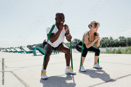 Jamaican man in white t-shirt and black shorts with elastic tape doing squats with women in grey top and black pants on fresh air and sunny weather