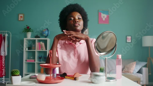 Portrait of young Afro-American woman sitting at beauty table in living room, in pink T-shirt and big earrings, looking at mirror and smiling, enjoying her beauty, Slow motion.