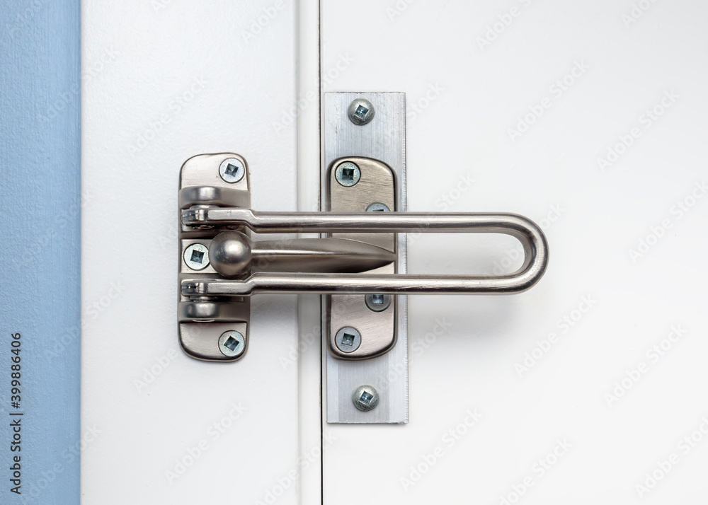 Door lock guard installed on white closed door, front view. Also known as hinged bar lock or security door guard. Home safety hardware limiting door openings in residences. Selective focus. Door lock guard installed on white closed door, front view. Also known as hinged bar lock or security door guard. Home safety hardware limiting door openings in residences. Selective focus.