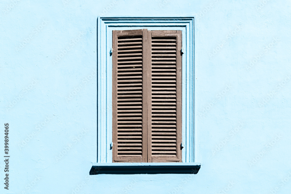 Gray shutters on the window against a blue colored wall. Old exterior in italian or greek village with closed window. Wooden vintage window with grey blinds