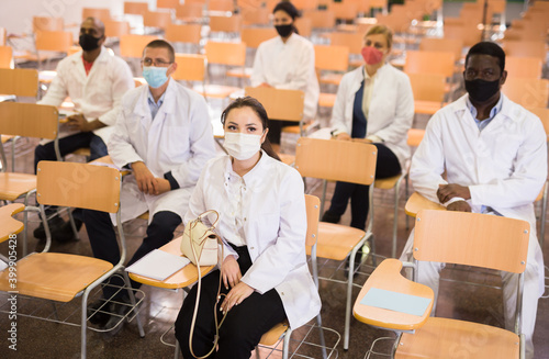 Group of people in white coats and protective masks sitting in conference room keeping distance at professional training for health workers. Precautions during mass events in coronavirus pandemic