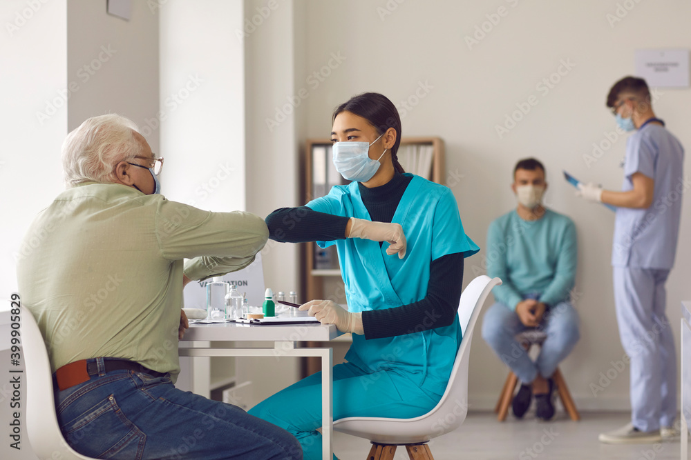 Fototapeta premium Young nurse in face mask bumping elbows with senior man before giving him vaccine