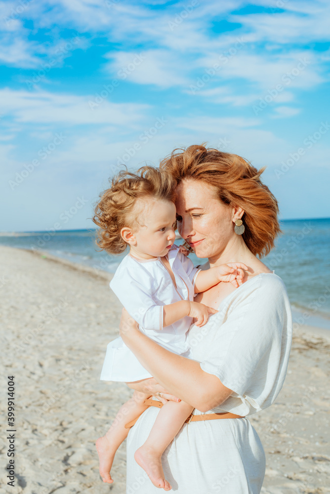 A young mother with red curly hair with her little daughter, a toddler in light clothes in the summer on the seashore, enjoy the rest. Happy family and children. Summer vacation and tourism

