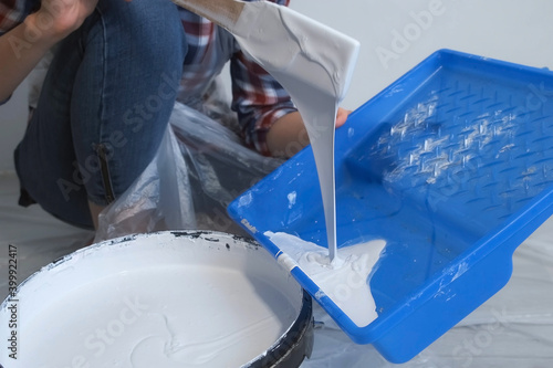 Woman painter is pouring paint in a tray for painting walls using wooden spatula, closeup view. DIY project, painting walls. Woman painter is doing renovation, finishing works at home by herself. photo