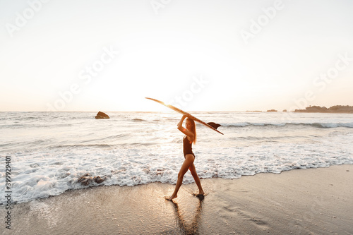Attractive young woman in black swimsuit is standing on beach with surfboard in hands, watch the sunset