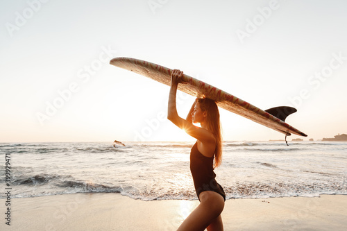 Photography Attractive young woman in black swimsuit is standing on beach with  surfboard in