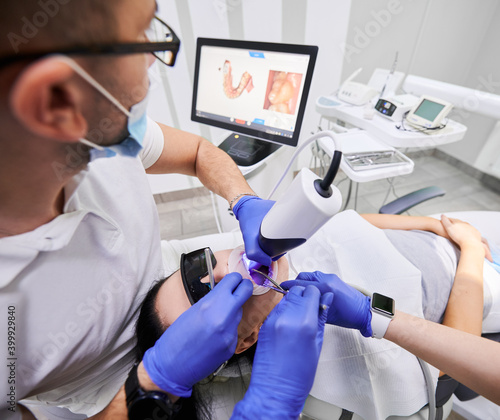 Dentist and his assistant scanning patient s teeth with modern scanning machine. Digital print of patient s teeth is on big screen. Modern high precision technologies. Concept of modern dentistry
