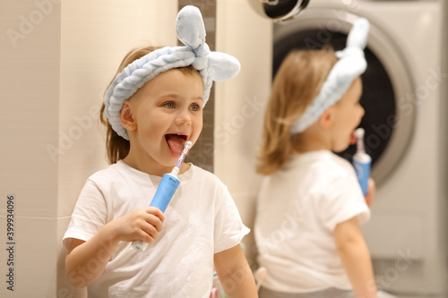 Adorible happy little girl in blue headbend holds toothbrush in bathroom. Beige background. Emotional impressed little girl opening mouth widely, having surprised facial expression. Hygiene concept. photo