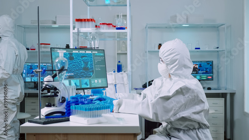 Woman biochemist in coverall checking manifestations of virus working on comuper in equipped lab. Team of doctors examining vaccine evolution using high tech researching diagnosis against covid19