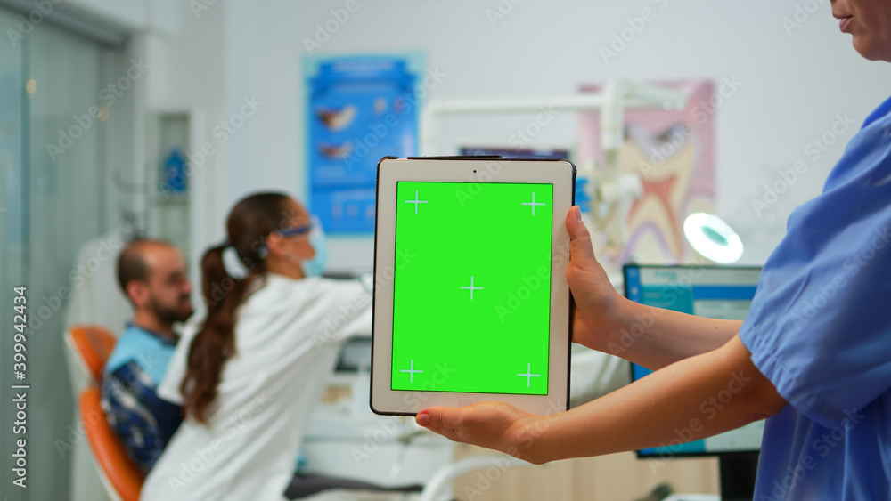 Close up of tablet with green screen display held by dental assistant standing in stomatologic room, while doctor examining patient in background. Using monitor with chroma key izolated pc key mockup Close up of tablet with green screen display held by dental assistant standing in stomatologic room, while doctor examining patient in background. Using monitor with chroma key izolated pc key mockup
