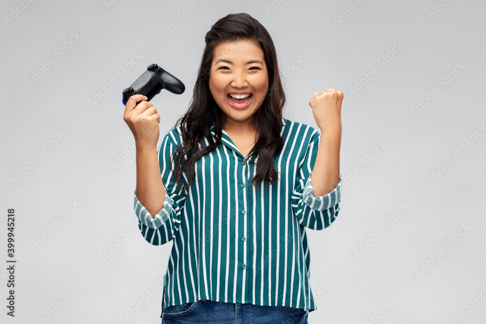 Fototapeta premium people, leisure and gaming concept - happy asian young woman playing video game with gamepad celebrating success over grey background