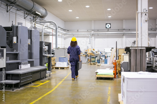 African American female industrial employee in hardhat and overall walking on plant floor. Back view. Work in factory or labor concept