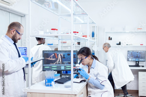 Assistant doing research using microscope and scientist taking notes on tablet pc. Team of researchers doing pharmacology engineering in sterile laboratory for healthcare industry with african