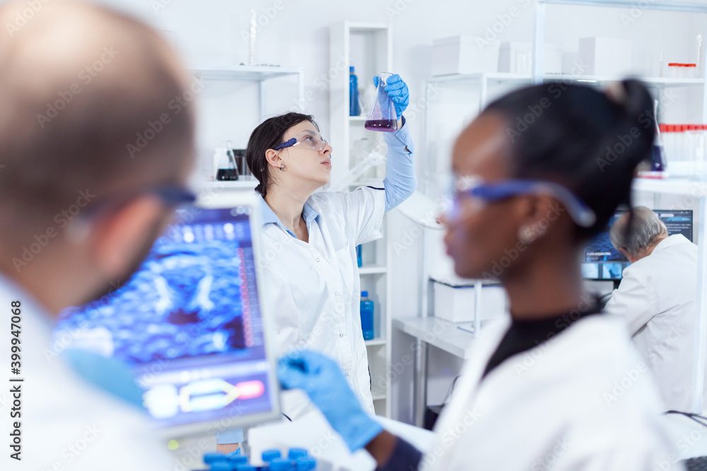 Female scientist holds flask with cure for vaccine during global pandemic. Multiethnic team of medical researchers working together in sterile lab wearing protection glasses and gloves. Female scientist holds flask with cure for vaccine during global pandemic. Multiethnic team of medical researchers working together in sterile lab wearing protection glasses and gloves.