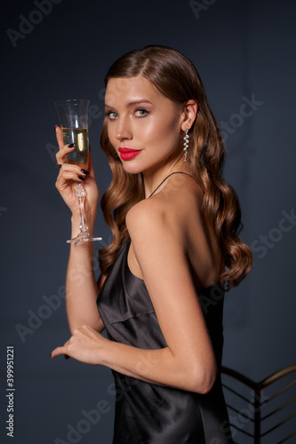 Portrait of elegant lady in black silk evening dress in dark interior. Beautiful caucasian woman with makeup and long wavy hair holding glass of white sparkling wine