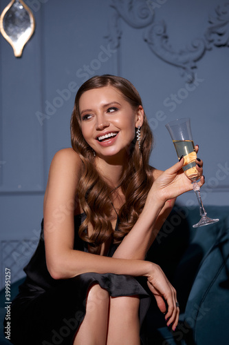 Elegant woman with perfect makeup and wavy brunette hair in long black silk evening dress sitting at sofa with glass of white sparkling wine in luxury stylish interior