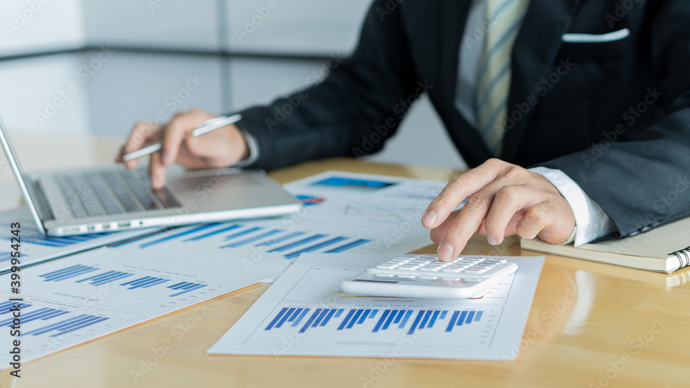 The hand that presses the calculator and holds the pen of a businessman working with a laptop with a graph is placed on a table with a blurred background. The hand that presses the calculator and holds the pen of a businessman working with a laptop with a graph is placed on a table with a blurred background.