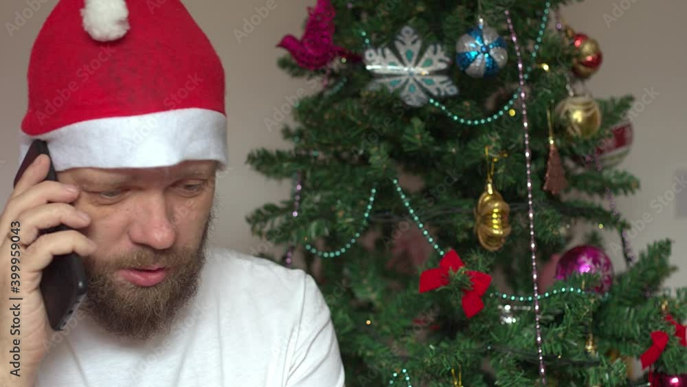 a cheerful man in a Santa Claus hat wishes his friends and family a merry Christmas using a video call, a family holiday online, a social distance during quarantine and lockdown a cheerful man in a Santa Claus hat wishes his friends and family a merry Christmas using a video call, a family holiday online, a social distance during quarantine and lockdown